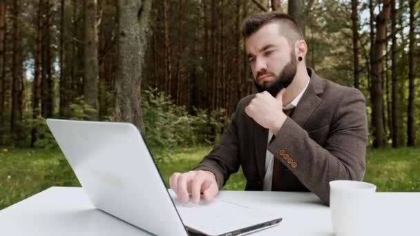 Giovane uomo d'affari attraente in abito marrone e cravatta si siede alla scrivania e lavora sul computer all'aperto. Alberi verdi, natura e parco sullo sfondo . — Video Stock
