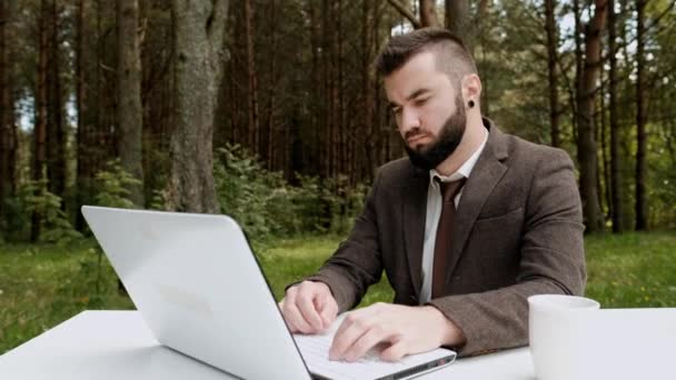 茶色のスーツとネクタイの若い魅力的な男性ビジネスマンは、机の上に座って、屋外でコンピュータ上で動作します。緑の木々、自然、公園を背景に. — ストック動画