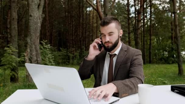 Joven atractivo hombre de negocios en traje marrón y corbata se sienta en el escritorio y trabaja en la computadora al aire libre. Árboles verdes, naturaleza y parque en el fondo . — Vídeos de Stock