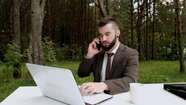 Fiatal, vonzó férfi üzletember barna öltönyben és nyakkendőben ül az asztalnál és számítógépen dolgozik a szabadban. Zöld fák, természet és park a háttérben. — Stock videók