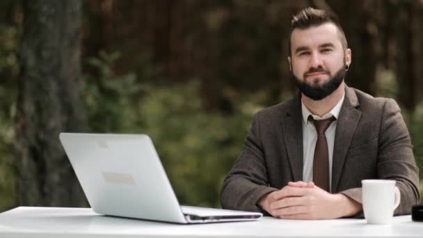 Mladý atraktivní mužský obchodník v hnědém obleku a kravatě sedí za stolem a pracuje na počítači venku. Zelené stromy, příroda a park na pozadí. — Stock video