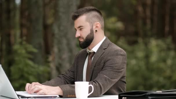 Young attractive male businessman in brown suit and tie sits at desk and works on computer outdoors. Green trees, nature and park on background. — Stock Video