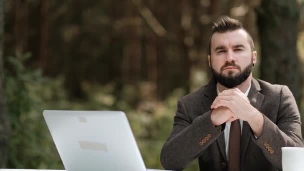 Joven atractivo hombre de negocios en traje marrón y corbata se sienta en el escritorio y trabaja en la computadora al aire libre. Árboles verdes, naturaleza y parque en el fondo . — Vídeo de stock