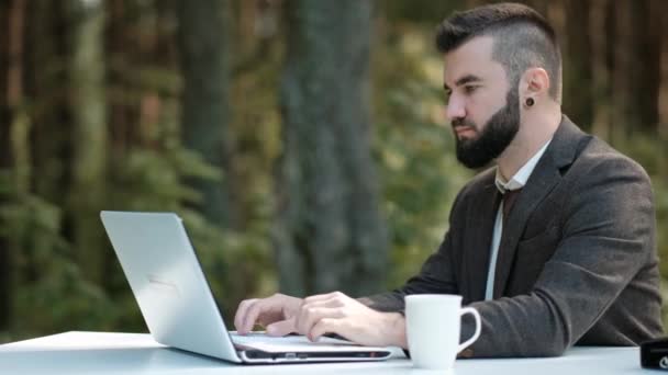 Jonge aantrekkelijke mannelijke zakenman in bruin pak en stropdas zit aan het bureau en werkt op de computer buiten. Groene bomen, natuur en park op achtergrond. — Stockvideo