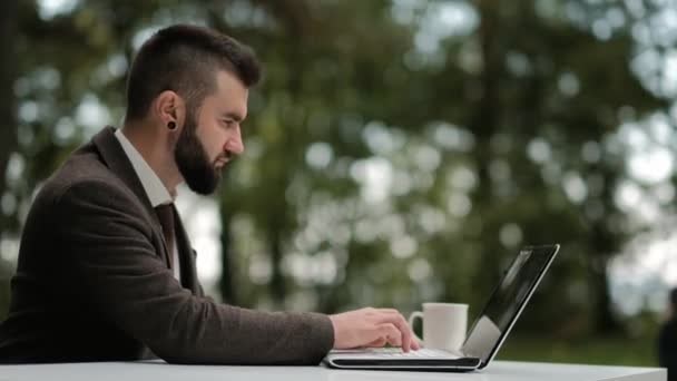 Joven y atractivo hombre de negocios barbudo sentado en un escritorio con una computadora portátil y trabajando remotamente al aire libre . — Vídeo de stock