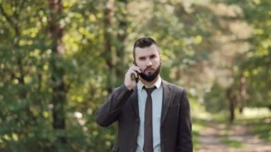 Kahverengi takım elbiseli genç ve çekici erkek işadamı yeşil bir yaz parkında yürüyor ve telefonda konuşuyor..