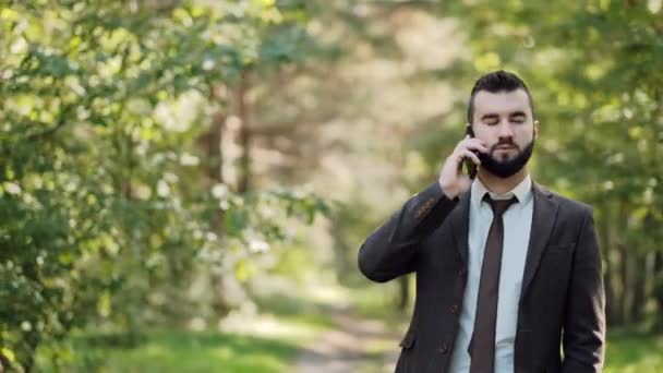 Jonge aantrekkelijke mannelijke zakenman in een bruin pak loopt in een groen zomerpark en spreekt aan de telefoon. — Stockvideo