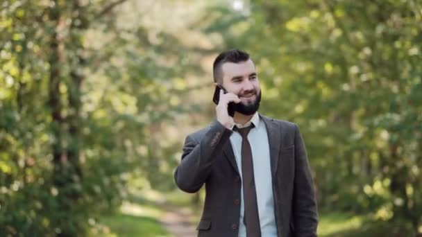 Mladý atraktivní muž podnikatel v hnědém obleku procházky v zeleném letním parku a mluví po telefonu. — Stock video