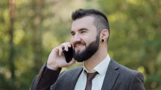 Joven atractivo hombre de negocios en un traje marrón camina en un verde parque de verano y habla por teléfono . — Vídeos de Stock
