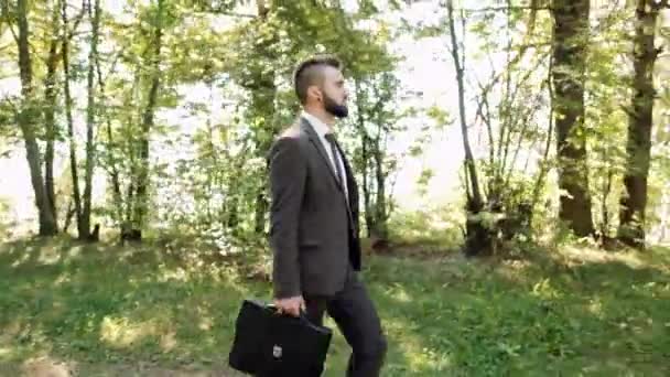 Jeune homme d'affaires attrayant dans un costume brun avec une mallette marche dans un parc verdoyant . — Video