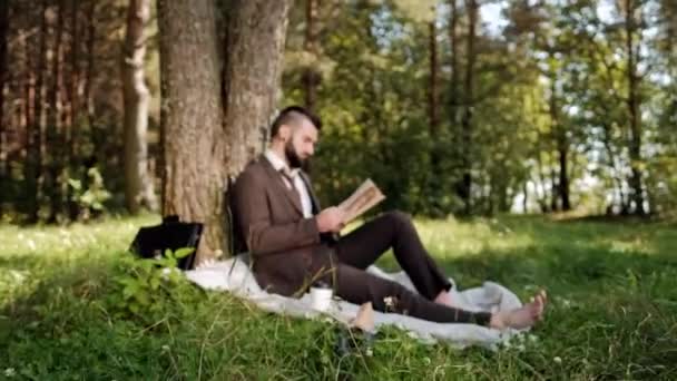 Giovane uomo d'affari attraente in un abito marrone con una valigetta seduta sull'erba sotto un albero in un parco, a riposo, mangiare un panino e bere caffè . — Video Stock