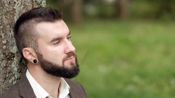 Joven atractivo hombre de negocios en traje marrón con un maletín sentado en la hierba debajo de un árbol en un parque, descansando, comiendo un sándwich y tomando café . — Vídeos de Stock