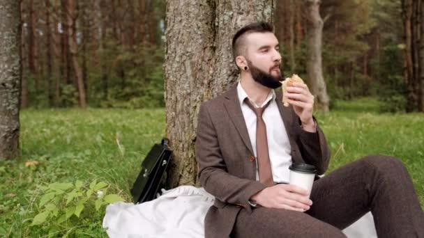公園の木の下の草の上に座っているブリーフケース付きの茶色のスーツを着た若い魅力的な男性実業家、休憩、サンドイッチを食べてコーヒーを飲む. — ストック動画