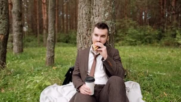 Jovem atraente homem de negócios em um terno marrom com uma pasta sentada na grama sob uma árvore em um parque, descansando, comendo um sanduíche e bebendo café . — Vídeo de Stock