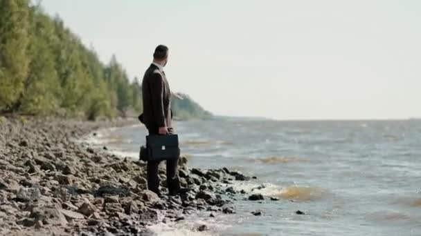 Jeune homme d'affaires attrayant dans un costume brun avec une mallette se tient sur le bord de la mer . — Video