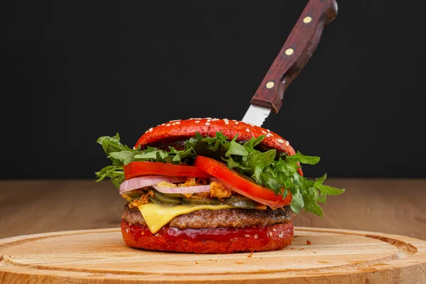 Delicioso hambúrguer picante vermelho com carne de boi, pão e feitiçaria e pimenta em uma tábua de madeira com uma faca picada em um fundo preto . — Fotografia de Stock