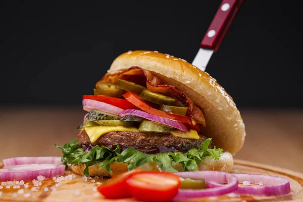 Delicious juicy burger with beef patty, bun and vegetables on a serving wooden board with a knife stuck. Food delivery. Fast food restaurant.