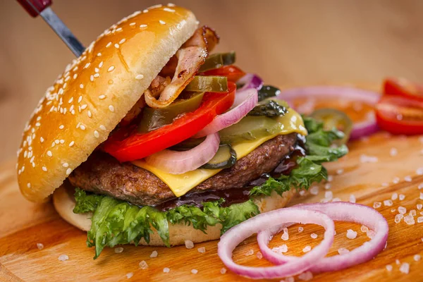 Delicioso hambúrguer suculento com carne bovina, pão e legumes em uma tábua de madeira com uma faca presa. Entrega de comida. Restaurante fast food . — Fotografia de Stock