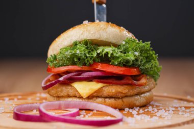 Siyah arka planda saplanmış bir bıçakla servis edilmiş ahşap bir tahtada lezzetli sulu bir hamburger. Biftekler, sebzeli çörekler ve doğranmış soğan..