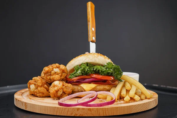 Combo set of fast food. Juicy hamburger, French fries, chicken legs and sauce. Served wooden board and cutlery on a black background. Fast food restaurant. Food delivery.