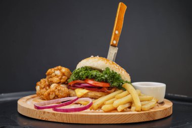 Fast food seti. Sulu hamburger, patates kızartması, tavuk bacağı ve sos. Siyah arka planda ahşap tahta ve çatal bıçak servisi yapıyordu. Fast food restoranı. Yemek teslimatı.