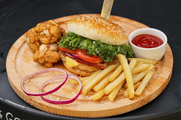 Combo set of fast food. Juicy hamburger, French fries, chicken legs and sauce. Served wooden board and cutlery on a black background. Fast food restaurant. Food delivery.