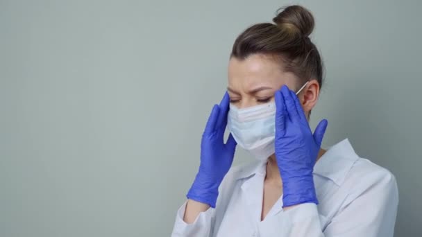 Портрет молодой женщины-врача в белом халате, маске и перчатках на сером фоне. Уставший медицинский работник болен. . — стоковое видео