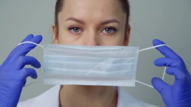 Beyaz cüppeli ve eldivenli genç bir kadın doktor yüzüne koruyucu bir maske takıyor..