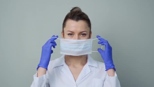 Uma jovem médica em um manto branco e luvas coloca uma máscara protetora em seu rosto . — Vídeo de Stock