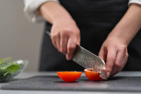 Una cuoca in uniforme bianca e un grembiule nero nella cucina del ristorante. Il cuoco taglia le verdure con un grande coltello da cucina. — Foto Stock