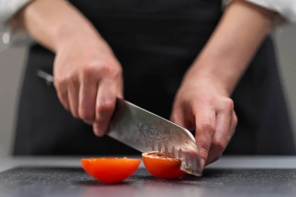 Una cuoca in uniforme bianca e un grembiule nero nella cucina del ristorante. Il cuoco taglia le verdure con un grande coltello da cucina. — Foto Stock