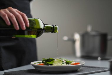 Beyaz üniformalı bir kadın şef ve restoran mutfağında siyah önlük. Yemek pişirmek. Aşçı yeşil bir şişeden zeytinyağı döker..