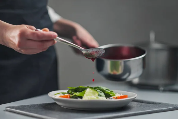 Una cuoca in uniforme bianca e un grembiule nero nella cucina del ristorante. Il cuoco versa salsa di insalata di mirtilli rossi. Fare e decorare il cibo. — Foto Stock