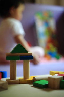 Arka planda oyuncak bloğu oynayan bulanık bir bebek. Renkli ahşap bloklardan yapılmış kaleye odaklanmış. Bebek oynayarak öğreniyor. Beyin ve kas gelişimi genç ve bebek safhasında.