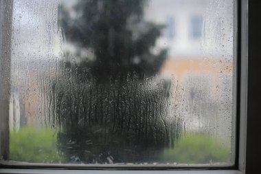 Pencere yağmurdan sırılsıklam olmuş. Su damlası şeffaf cam pencereye takıldı. Caddenin diğer tarafındaki evin bulanık görüntüsü. Yağmur ve fırtına mevsiminde evde kal.