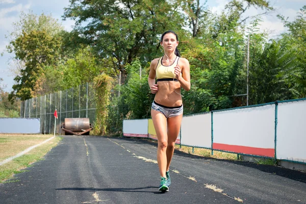 Chica delgada con una figura deportiva en pantalones cortos corriendo alrededor del estadio escuchando música —  Fotos de Stock