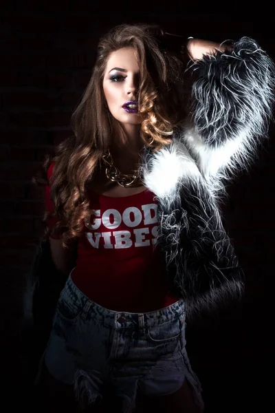 Primer plano, muy lindo sexy chica joven o mujer con pelo largo rizado y maquillaje de moda en pantalones cortos vaqueros y camisetas rojas, en la oscuridad sobre un fondo de una pared de ladrillo, espacio de copia para el texto — Foto de Stock