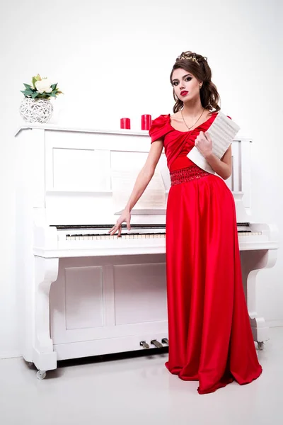 Элегантная женщина в длинном красном платье стоит у фортепиано с неопознанными нотами в руках — стоковое фото