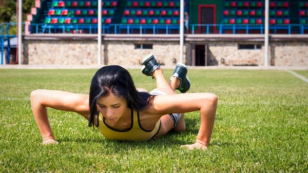 Стройная девушка со спортивной фигурой в коротких шортах делает упражнения на стадионе, на газоне — стоковое фото