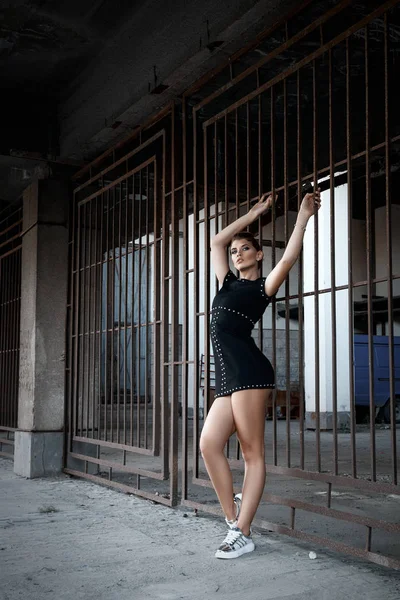 Красивая молодая девушка с красивым макияжем, в коротком черном платье, прижата к решетчатым воротам — стоковое фото