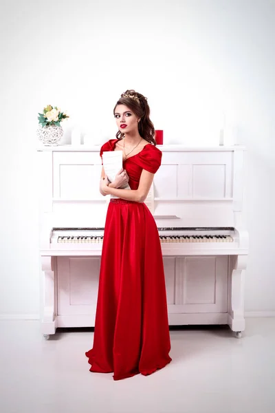 Elegante mujer con un vestido rojo largo sostiene notas no reconocidas, de pie al piano en una habitación blanca —  Fotos de Stock