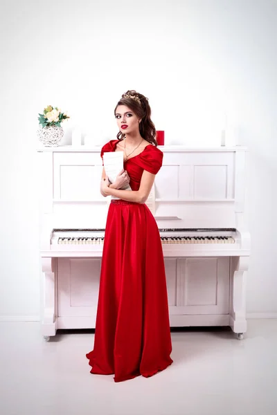 Elegant woman in a long red dress holds unrecognized notes, standing at the piano in a white room Stock Picture