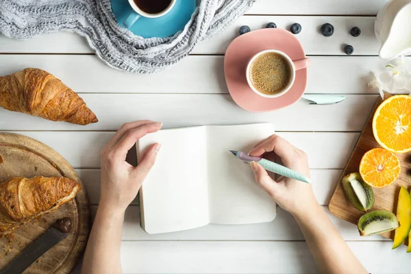 Mladá žena má na snídani s čerstvými croissanty, kávu a ovoce a její ruce, kreslení nebo psaní perem inkoustu v otevřít poznámkový blok na bílý dřevěný stůl. Stock Obrázky