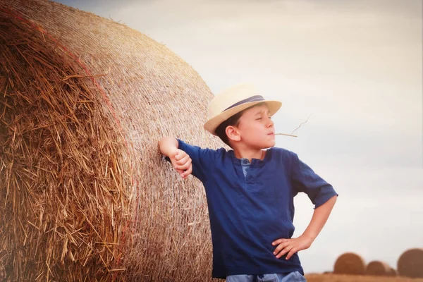 Young cute country boy in hat near haystacks at sunset in summer.a summer concept, a vacation. A cute boy is sitting on a haystac — Stock Photo, Image