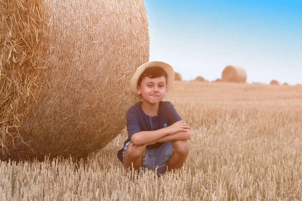 Fiatal aranyos vidéki fiú, kalapban közelében haystacks napnyugtakor a summer.a nyári koncepció, a nyaralás. Aranyos fiú ül egy haystac — Stock Fotó