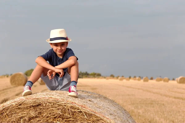 Симпатичный мальчик сидит на стоге сена. Активное времяпровождение на свежем воздухе с детьми в теплый летний день. . — стоковое фото