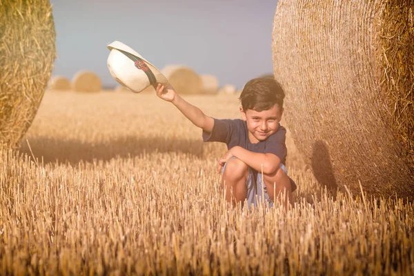 Молодой милый деревенский мальчик в шляпе возле стога сена на закате летом. Летняя концепция, отдых. Активный отдых на свежем воздухе с детьми в теплый летний день . — стоковое фото