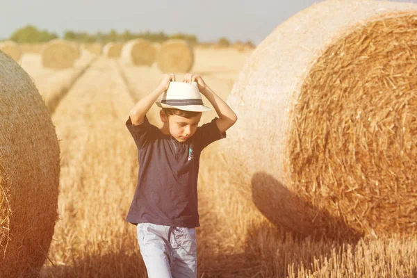 Молодий симпатичний сільський хлопчик у капелюсі біля сінокосів на заході сонця влітку. Літня концепція, відпустка. Активний відпочинок на відкритому повітрі з дітьми в теплий літній день . — стокове фото