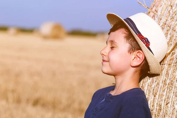 Young cute country boy in hat near haystacks at sunset in summer. Summer concept, a vacation.Active outdoors leisure with children on warm summer day. — Stock Photo, Image