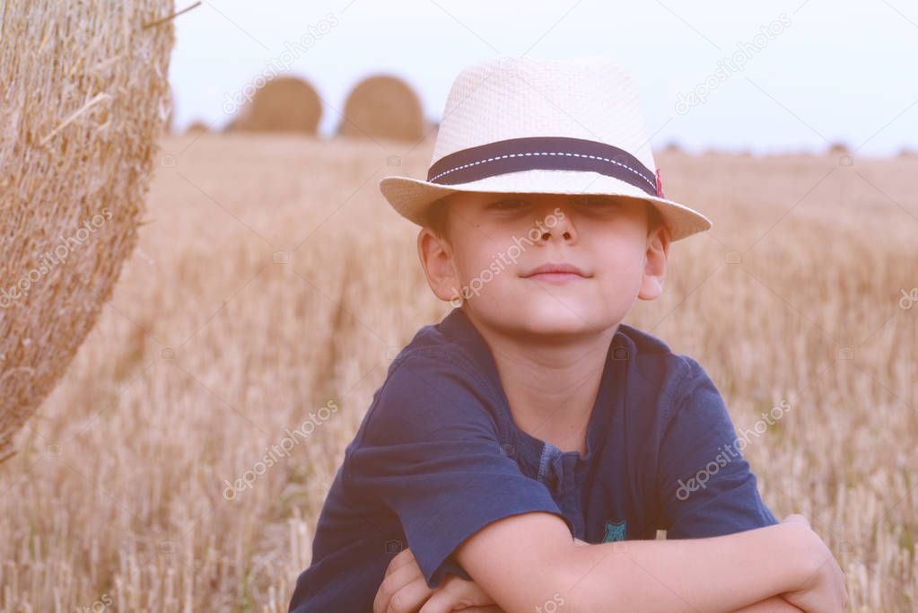 Young cute country boy in hat near haystacks at sunset in summer. Summer concept, a vacation.Active outdoors leisure with children on warm summer day.